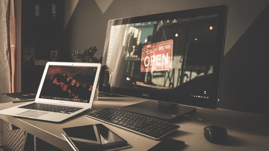 A photograph of a dramatically lit room witrh two computers. The reflection in one of the monitors has a "come in, were open" sign