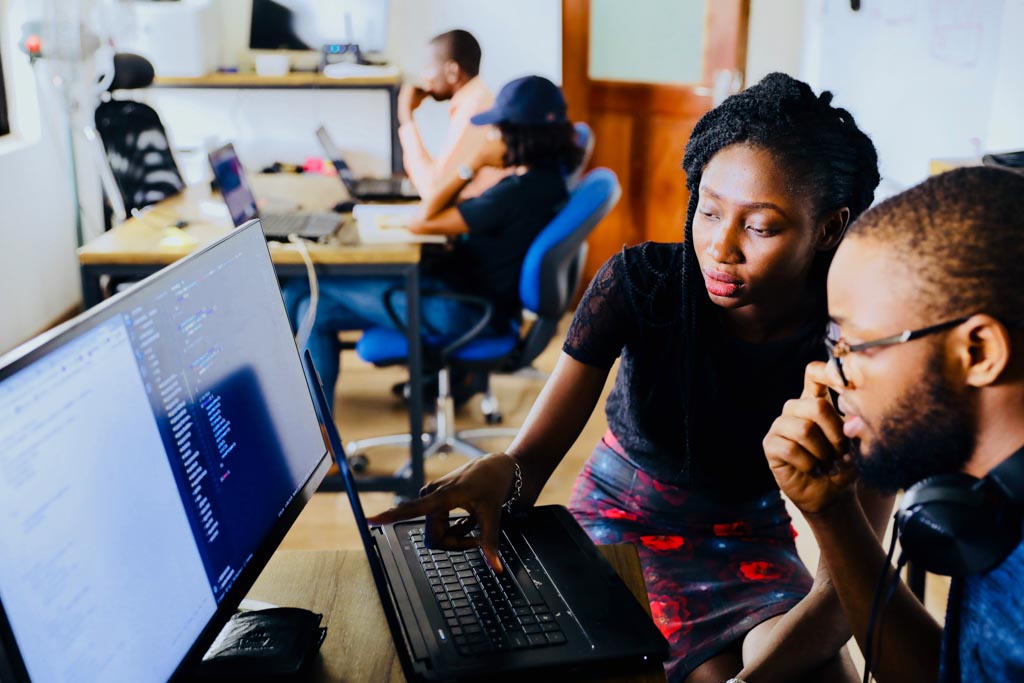 A woman and a man look at the same computer screen. They are in a computer lab with other people in the background