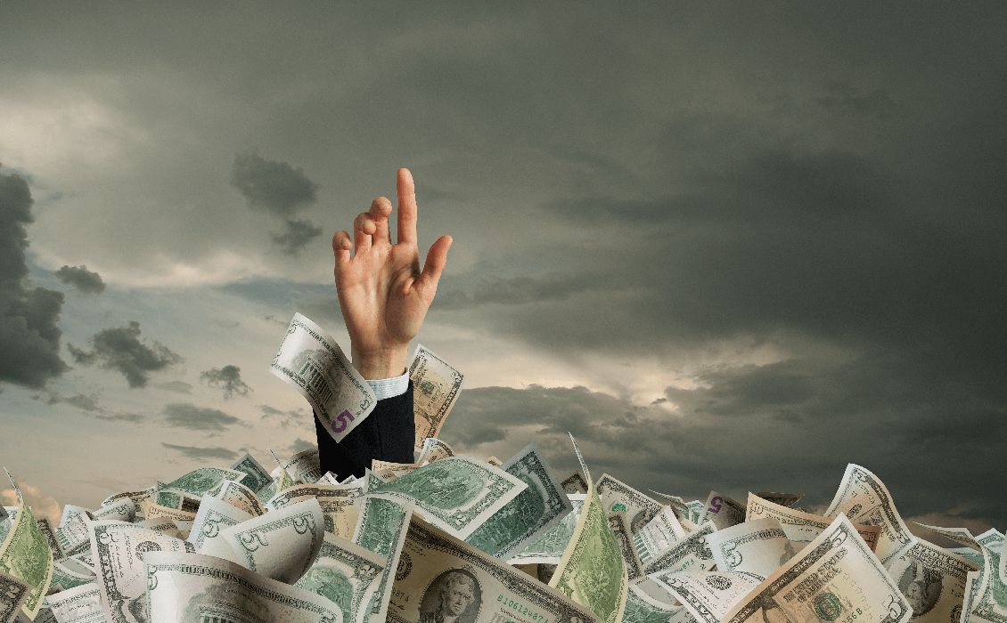 Hand reaching out of piles of US dollars, as if drowning in money.
